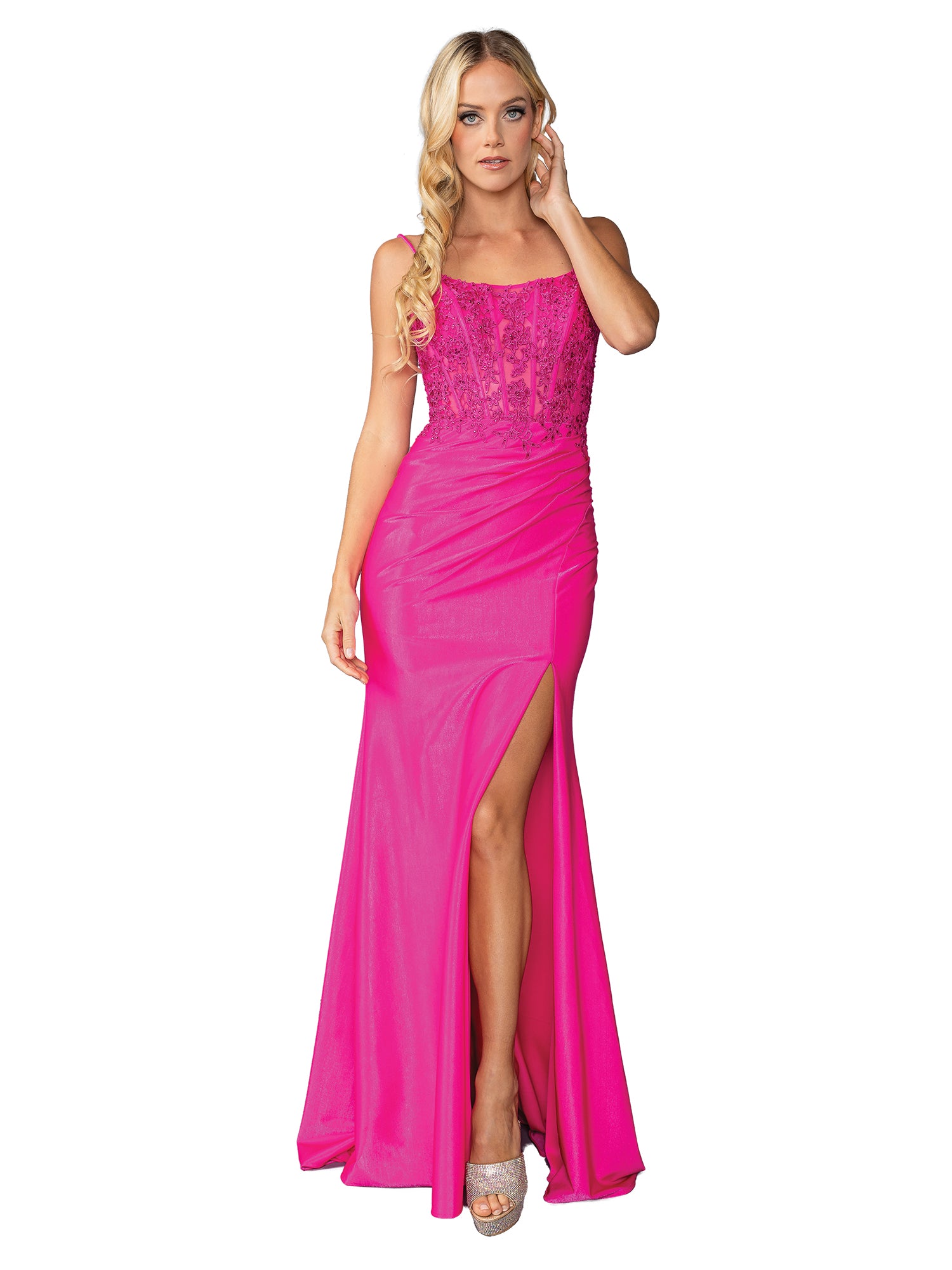 A woman wearing the Lace-Up Long Fitted Prom Dress: Dancing Queen 4434 in bright pink with a sheer corset bodice, side slit, and silver heels stands against a white background.